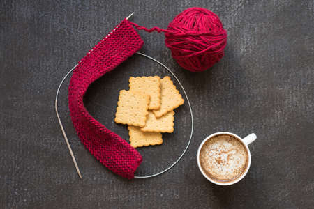 Knitting with red yarn and needles, a cup of coffee and crackers on a dark painted grunge background.の写真素材