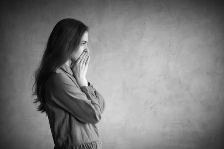 Woman is standing hear the grunge wall. She is sad and depressed, covering her mouth with hands. Retouched and vignette is added.の写真素材
