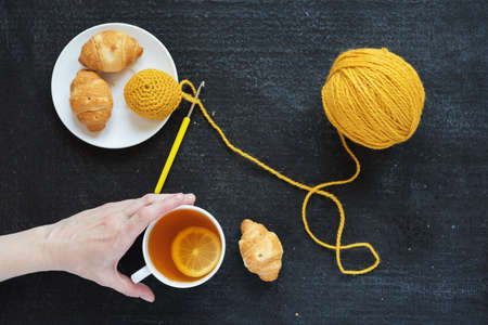 Lemon tea, yellow crocheting and croissants on a plate. Female hand is taking a cup of tea.の写真素材