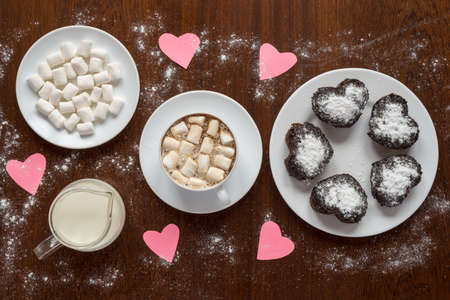 Paper hearts on a coffee table on wooden background.There are also a cup of coffee, a plate with cup cakes, a saucer with marsh mallow and a glass jar with milk.の写真素材