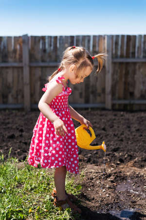 Little girl in a red spotted dress with a yellow toy watering-can in the garden. The day is sunny and the shadow are deep. She is watering the garden.の写真素材