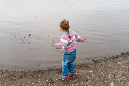 Little three-year-old girl in a sweater and jeans throwing little stones into the river.の写真素材