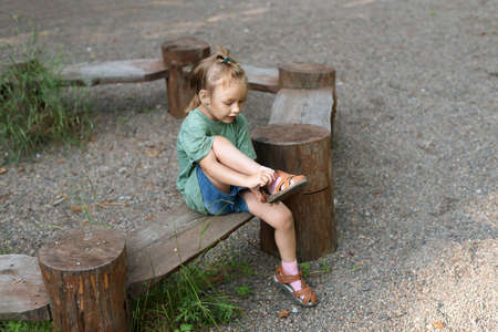 Little girl putting her sandal on. She is sitting on a wooden round bench in a summer forest.の写真素材