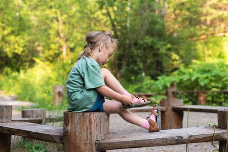 Little girl putting her sandal on. She is sitting on a wooden round bench in a summer forest.の写真素材
