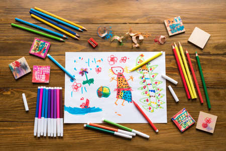Crayons, felt-tip pens and a child's drawing on a wooden table.
