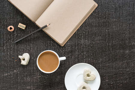 Cup of coffee, diary with empty brown pages, a black pencil with a sharpener and a plate with three heart-shaped cookies on a black shabby background.の写真素材