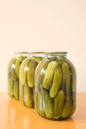 Three closed glass transparent jars with fresh pickled cucumbers placed on a wooden table on white background, homemade preserved vegetables.の写真素材