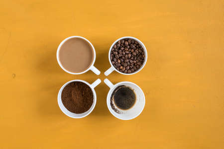 Four different cups on a yellow background. First with coffee beans, second with ground coffee, third with milk coffee and forth with just drunk coffee. Shot overhead.の写真素材