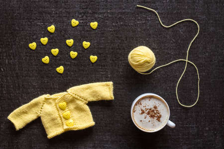 Cup of coffee, yellow knitted sweater for a doll and a heart-shaped buttons on a grunge black background.の写真素材