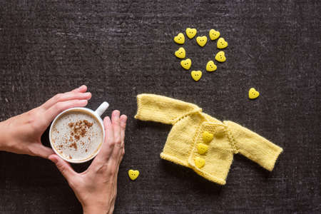 Cup of coffee, yellow knitted sweater for a doll and a heart made of buttons on a grunge black background. Female hands holding a cup.の写真素材