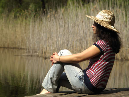Girl in straw hat sitting near lake shoreの写真素材