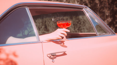 A woman's hand holds a glass with ice cubes and a red cocktail inside a pink vintage car.の素材