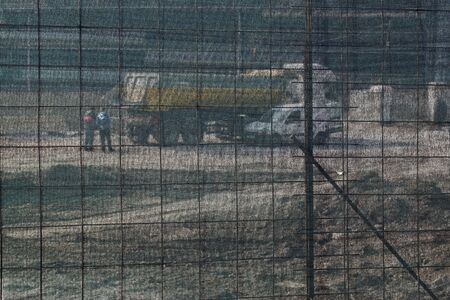 Unrecognizable, unidentifiable construction workers and a truck at  construction site, behind plastic safety protective net. Selective focus background.の写真素材