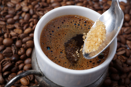 Close up of traditional homemade  Turkish coffee.
Adding spoonful of granulated crystal brown sugar.
Strong unfiltered coffee in white porcelain cup. 
Coffee beans background.
Tradition concept. の写真素材