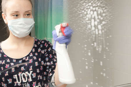 cute young girl maid wearing a medical mask sprinkles cleaning agent on the bathroom mirror during the coronavirus pandemicの写真素材