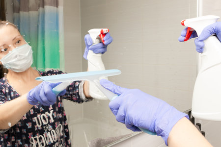 cute young girl maid in medical mask cleans bathroom mirror during coronavirus pandemic