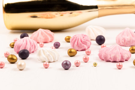 golden gift bottle of champagne, multicolored dragee candies and meringue cake on white wooden table