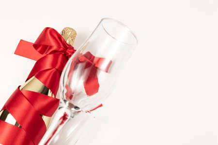 empty wine glass in the foreground against the background of a bottle of champagne wrapped with a red silk ribbon on a white backgroundの写真素材