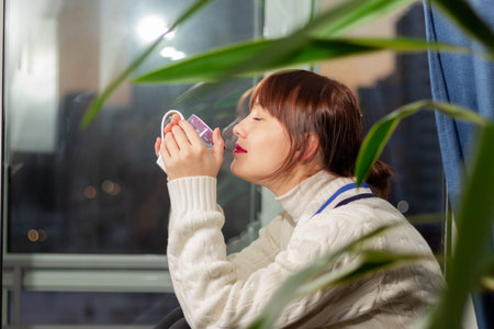 a young pretty girl in a warm sweater sits on the windowsill by the window and holds a mug of coffeeの写真素材