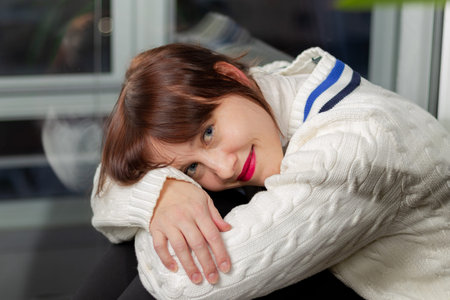young pretty girl in a white sweater sitting on the windowsill at home by the window dreaming