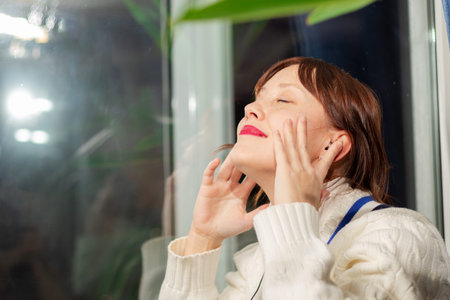 young pretty girl in headphones listens to music with pleasure while sitting on the windowsill at...