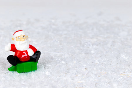 toy santa claus in a red jacket sits on a green chest with gifts on a background of white snowの写真素材