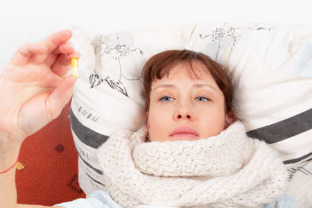 a young pretty girl wrapped in a scarf is ill at home, lies in bed and holds a capsule with medicine in her handの写真素材