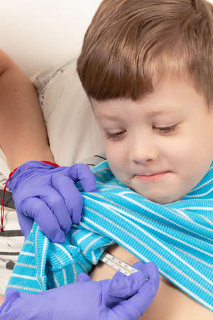 a sick child a boy in a blue jacket lies at home in bed and measures the temperature with a thermometerの写真素材