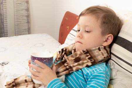 sick child boy in a blue sweater wrapped in a warm scarf in bed drinking ginger tea with honeyの写真素材