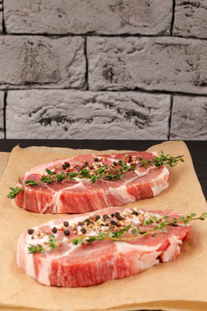 fresh raw marbled meat pork steak with thyme and seasoning on brown parchment paper on a black table against a gray brick wall backgroundの写真素材