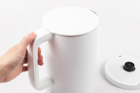 a female hand holds a white electric kettle by the handle on a white wooden table backgroundの写真素材