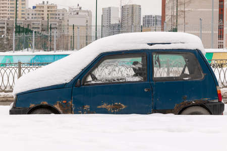 old blue abandoned broken rusty car parked by the road under a large layer of snow on a cold winter dayの写真素材