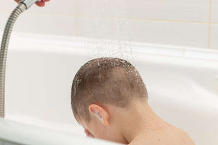 preschooler boy washes in a light white bathroom at home. water pours on top of his head from the shower headの写真素材