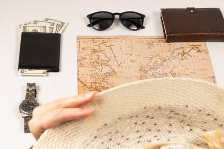 female hand holds a straw hat. on a white wooden table there is a world map, a black wallet with money, a diary, a watch and sunglasses. selective focusの写真素材