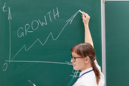young woman business coach in a white blouse and glasses at the chalkboard in the office draws a...