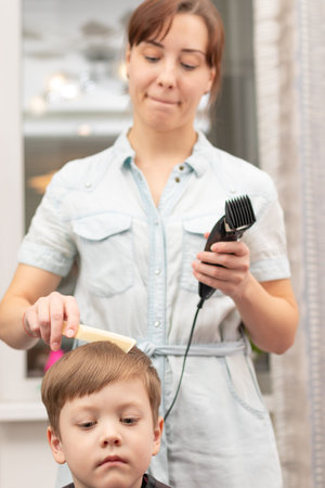 young mom hairdresser cuts her baby boy at home with hair clipper during quarantine. selective...