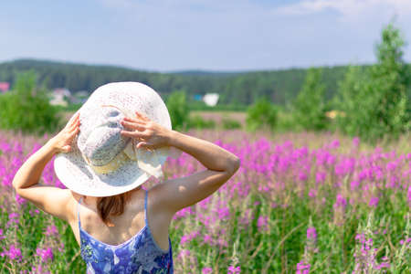 Young woman in a field of flowers in a straw hat against a background of green forest and blue sky on a hot summer day. Selective focusの写真素材