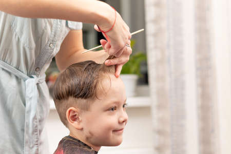caring female hands of a mother with a comb and scissors make a fashionable haircut for her son at home during the second lockdown. selective focusの写真素材