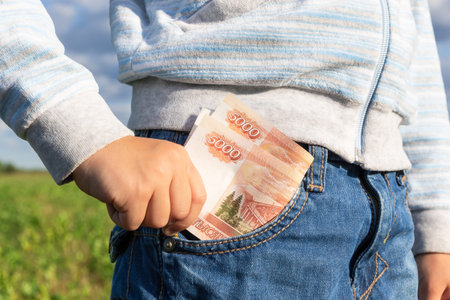 A child's hand takes out paper money rubles from a jeans pocket on a background of blue sky on a...