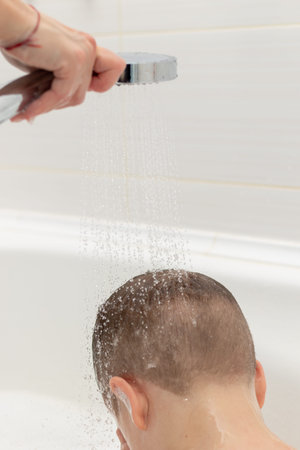 preschooler boy washes in a light white bathroom at home. water pours on top of his head from the...