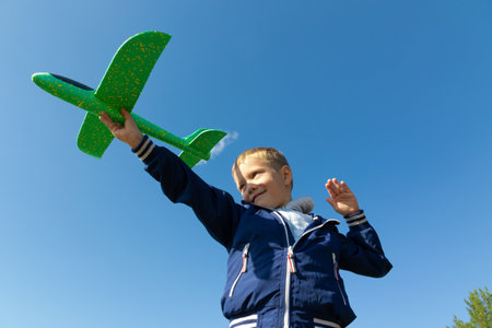 A six-year-old preschooler boy in a blue jacket launches a toy plane in nature against the background of a clear blue sky on a summer day. The bright sun is shining. Portraitの写真素材