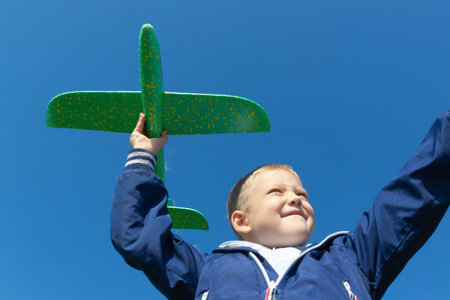 A six-year-old preschooler boy in a blue jacket launches a toy plane in nature against the...