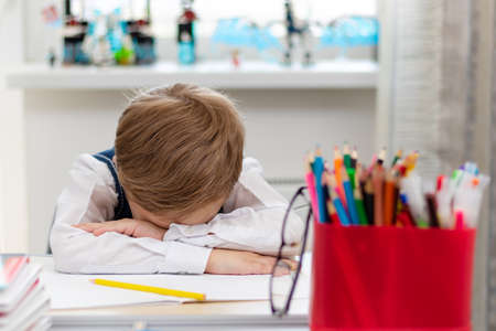 A cute first grader boy in a school uniform at home during a pandemic fell asleep doing homework at a desk with books and pencils. Selective focus. Close-up. Portraitの写真素材