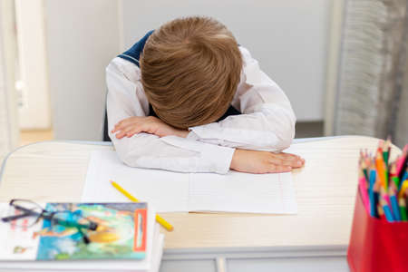 A cute first grader boy in a school uniform at home during a pandemic fell asleep doing homework at a desk with books and pencils. Selective focus. Close-up. Portraitの写真素材