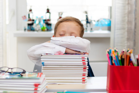 A cute first grader boy in a school uniform at home during a pandemic fell asleep doing homework at...