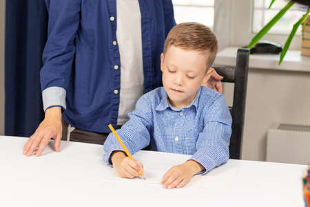 Cute seven year old baby boy doing homework at home with mom on a white wooden table during a pandemic. Selective focus. Close-up. Portraitの写真素材