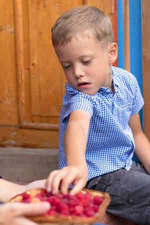 A six-year-old contented boy eats fresh fragrant raspberries in the village outdoors on the doorstep of the house. Selective focus. Portraitの写真素材