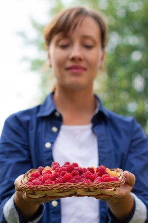 A beautiful young woman in a blue shirt holds a basket of fresh fragrant raspberries in her hands against a background of green foliage. Selective focus. Portraitの写真素材