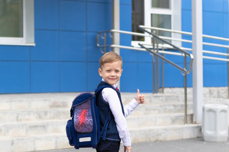 A cute first grader boy in a school uniform with a schoolbag goes to school on a sunny autumn day....