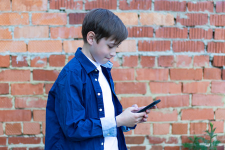 A teenage boy in a blue shirt with a smartphone in his hands communicates with friends on social networks against the background of a brick wall. Selective focusの写真素材